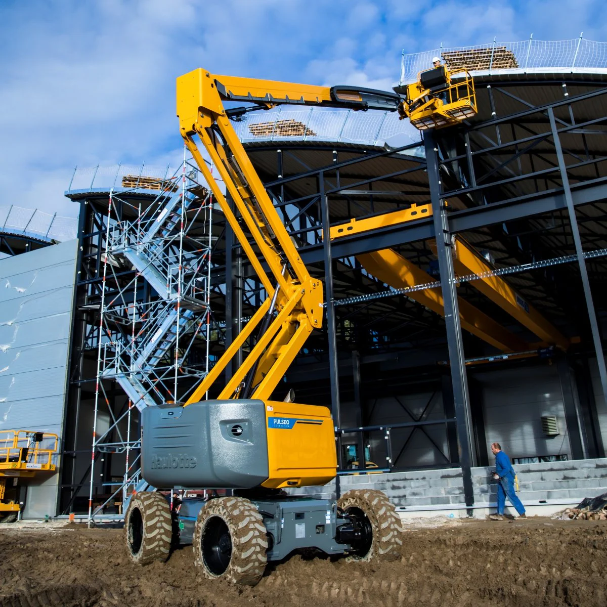 Large yellow articulated crane at a building under construction with scaffolding and a worker nearby.