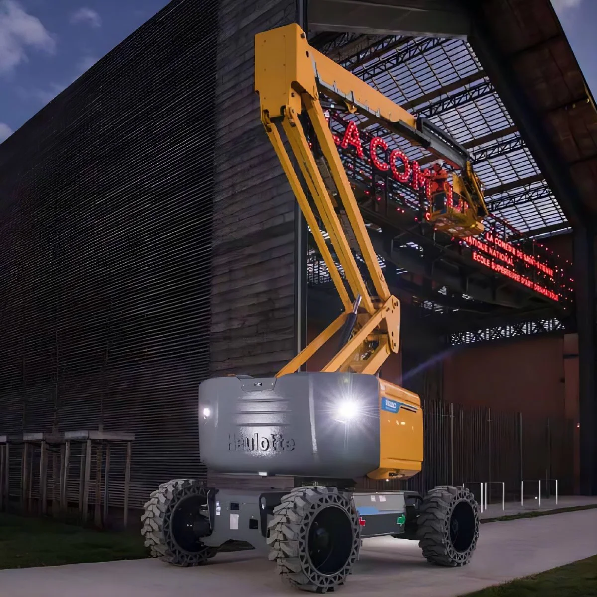 Telescopic boom lift with yellow arm and gray base parked beside a darkened building at dusk