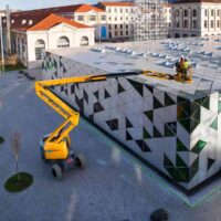 Two construction workers in safety vests on a yellow boom lift work along a geometric-patterned building facade.