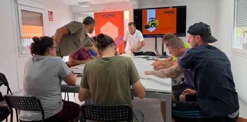 Instructor leads a workshop for a group around a table in a bright classroom; attendees listen and take notes as slides display on two screens.